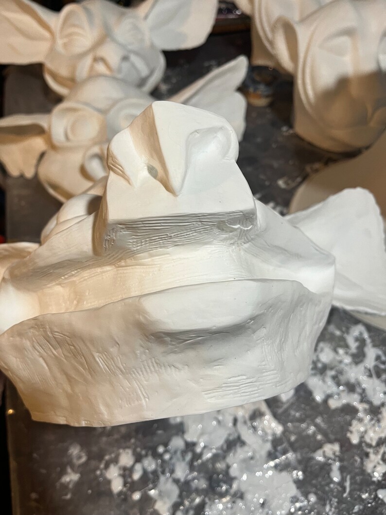 May include: A collection of unfinished, white plaster gargoyle sculptures. The sculptures feature detailed facial features, including prominent noses, open mouths, and textured surfaces. The gargoyles are in various stages of completion, suggesting a workshop or studio setting.