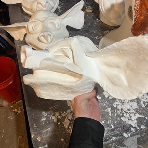 May include: A collection of unfinished, white plaster masks, including several goblin-like faces with large ears and expressive features. One mask is held by a person, showcasing the intricate details and textures of the artwork.