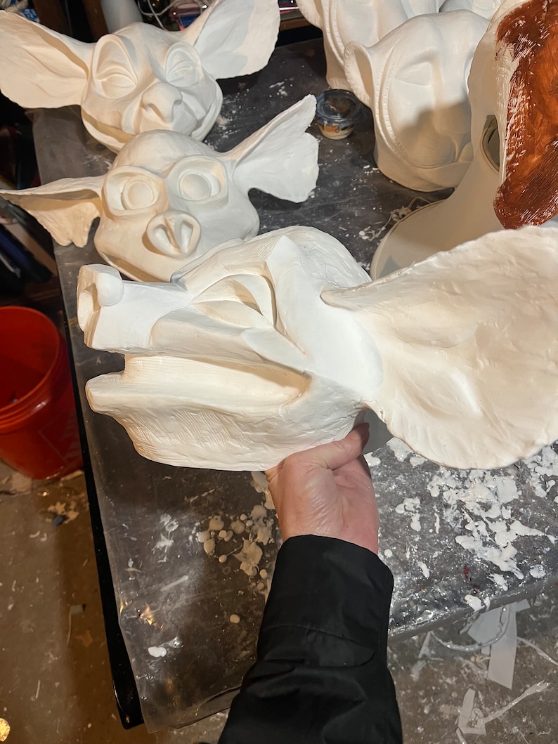 May include: A collection of unfinished, white plaster masks, including several goblin-like faces with large ears and expressive features. One mask is held by a person, showcasing the intricate details and textures of the artwork.