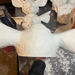 May include: Several white, unfinished masks with large ears and stylized facial features are arranged on a work surface. The masks appear to be made of a plaster or similar material. One mask is held by a person.