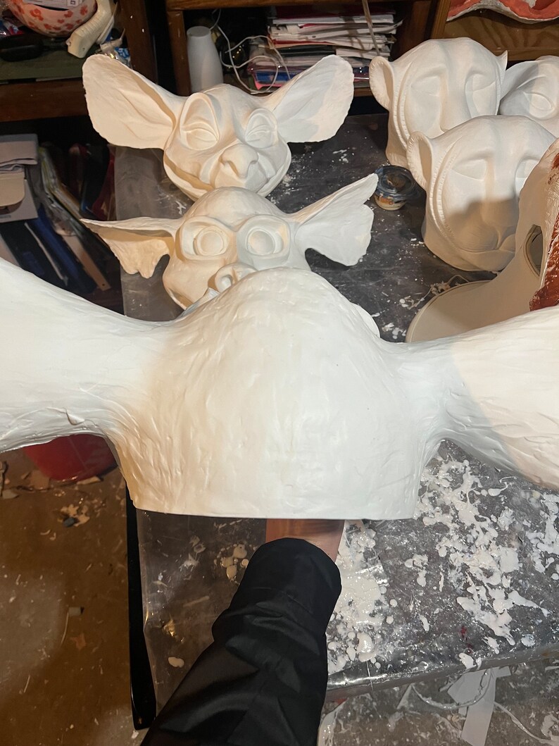 May include: Several white, unfinished masks with large ears and stylized facial features are arranged on a work surface. The masks appear to be made of a plaster or similar material. One mask is held by a person.