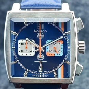 Op de afbeelding: Een vierkante polshorloge met een blauwe wijzerplaat en een blauwe leren band. De wijzerplaat heeft witte en oranje accenten, met de tekst "MONACO TAG HEUER" zichtbaar. Het horloge heeft zilverkleurige knoppen en een gesp.