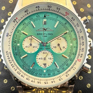 May include: A Breitling Navitimer watch with a metallic green dial, three cream-colored sub-dials, and a date window at the six o'clock position. The watch features a silver-tone bezel with a slide rule scale and a gold-tone metal link bracelet.