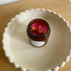 May include: A small, brown glass jar candle with pink wax and dried rose buds, in a cream-coloured, scalloped-edge ceramic dish. The candle has a white label with cursive text. The dish is on a light wood surface.