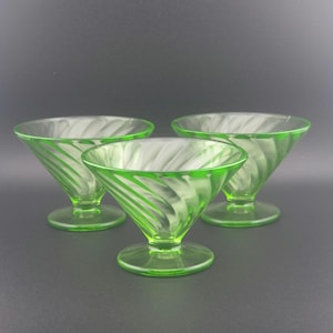 May include: Three vintage green glass dessert dishes. Each dish has a flared bowl with a spiral design and sits on a round base. The glass has a bright, translucent appearance, and the dishes are arranged against a dark background.
