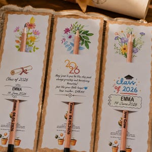 May include: Three rectangular paper cards with scalloped edges, each featuring a wooden pencil inserted through a slot. The cards display watercolor floral illustrations, the text Class of 2026, the name Emma, and the date 14 June 2026.