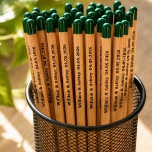 Puede incluir: Un conjunto de l&aacute;pices de madera clara con gomas de borrar de color verde oscuro en un portal&aacute;pices de malla met&aacute;lica negra. Cada l&aacute;piz tiene impreso el texto William & Ashley 04.05.2026 en tinta negra. Los l&aacute;pices est&aacute;n dispuestos verticalmente.