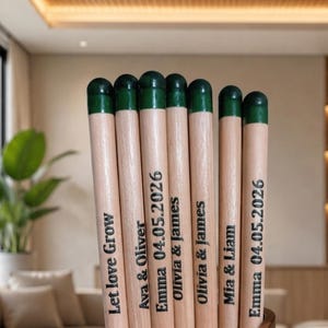Puede incluir: Una colecci&oacute;n de siete l&aacute;pices de madera clara con puntas pintadas en verde oscuro. Cada l&aacute;piz est&aacute; grabado con texto negro, incluyendo nombres como Ava & Oliver, Olivia & James y Mia & Liam, la fecha 04.05.2026 y la frase Let love Grow.