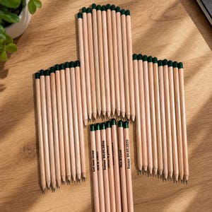 Puede incluir: Una colecci&oacute;n de l&aacute;pices de madera clara con gomas de borrar de color verde oscuro dispuestos en forma de cruz. Varios l&aacute;pices presentan texto impreso que incluye nombres como Emma, Mia y Liam, la fecha 04.05.2026 y la frase Let love grow.