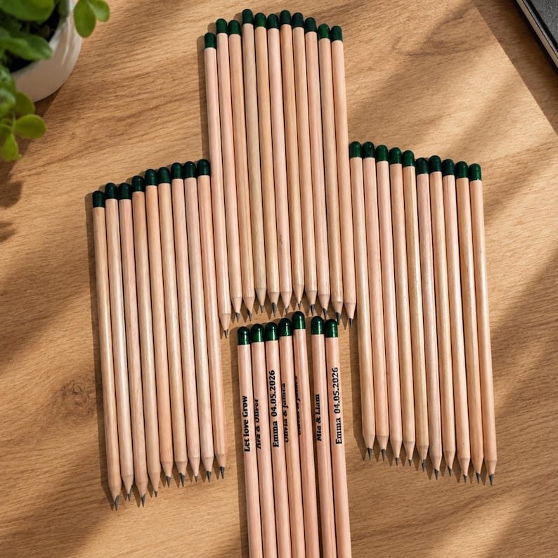 Puede incluir: Una colecci&oacute;n de l&aacute;pices de madera clara con gomas de borrar de color verde oscuro dispuestos en forma de cruz. Varios l&aacute;pices presentan texto impreso que incluye nombres como Emma, Mia y Liam, la fecha 04.05.2026 y la frase Let love grow.