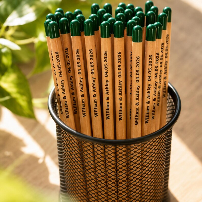 Puede incluir: Un conjunto de l&aacute;pices de madera clara con gomas de borrar de color verde oscuro en un portal&aacute;pices de malla met&aacute;lica negra. Cada l&aacute;piz tiene impreso el texto William & Ashley 04.05.2026 en tinta negra. Los l&aacute;pices est&aacute;n dispuestos verticalmente.