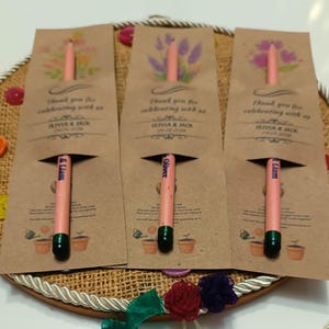 May include: Three seed-embedded pencils with green tips are partially visible in brown paper packaging. The packaging features floral illustrations and the text "Thank you for celebrating with us" along with names and a date. The pencils are a unique party favor.