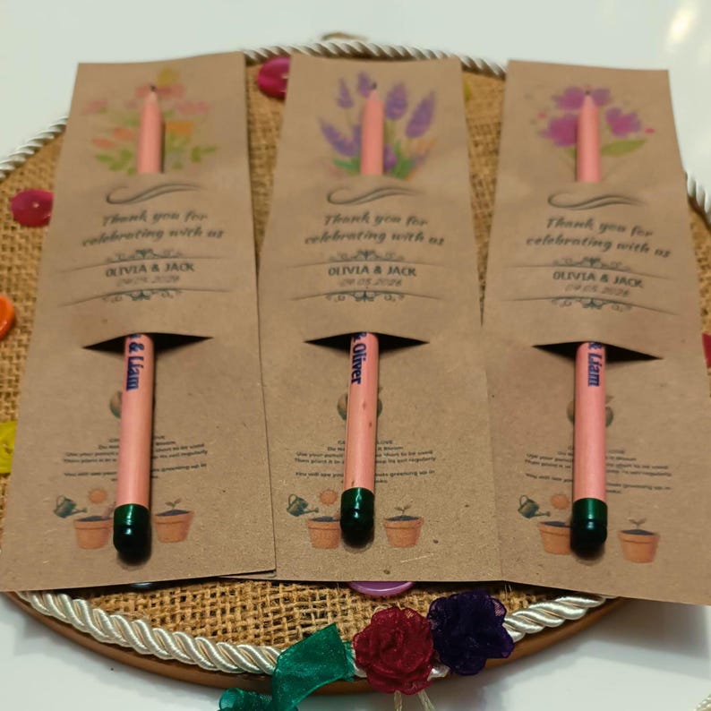 May include: Three seed-embedded pencils with green tips are partially visible in brown paper packaging. The packaging features floral illustrations and the text "Thank you for celebrating with us" along with names and a date. The pencils are a unique party favor.