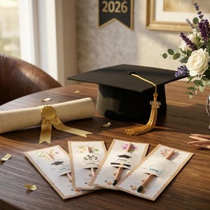 May include: A black graduation cap with a gold tassel featuring a charm that reads EMMA 2026, a rolled diploma tied with a gold ribbon, and four rectangular cards with floral designs, each holding a pencil and displaying graduation-themed text.
