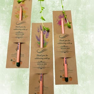 May include: Three kraft paper wedding favors with pencils. Each favor has a floral design and the text "Thank you for celebrating with us" and the names Olivia & Jack and the date 04.05.2026. The pencils are light pink with black tips.