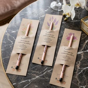 May include: Three pencils in kraft paper sleeves, each with a floral design and the text "Thank you for celebrating with us" and a date. The pencils are light pink with black tips. The sleeves are on a marble table.