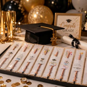 May include: A set of seven wooden pencils in individual clear plastic sleeves, each featuring a unique floral illustration and handwritten text. A black graduation cap with a gold 2026 tassel sits behind the pencils, alongside a framed sign.