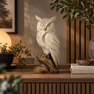 Può includere: Una statuetta decorativa a forma di gufo con corpo color crema caratterizzato da una texture venata e dettagli dipinti in oro su occhi, becco e piume delle ali. Il gufo è appollaiato su una base testurizzata di colore marrone scuro.