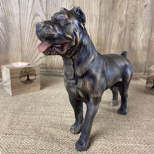 Könnte beinhalten: Eine stehende Hundefigur mit dunkelbronzenem Finish und goldfarbenen Akzenten. Die Skulptur zeigt einen muskulösen Hund mit kurzem Fell, spitzen Ohren und geöffnetem Maul, in dem eine rosa Zunge zu sehen ist. Die Figur ist realistisch gestaltet.