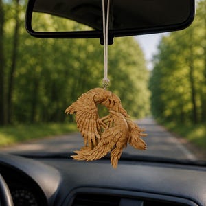 May include: A light brown wooden hanging ornament featuring two intricately carved ravens facing each other with their wings spread, surrounding a central triangular knotwork symbol. The ornament is suspended by a thin white cord from a rearview mirror.