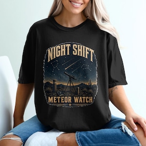 Puede incluir: Una camiseta negra con un estampado gráfico de un paisaje desértico nocturno con un telescopio, cactus y un cielo estrellado con meteoritos. El texto "NIGHT SHIFT" aparece en color beige sobre la escena y "METEOR WATCH" debajo de ella.