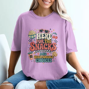 May include: A light purple short-sleeve t-shirt featuring a graphic print with the text "HERE FOR THE SNACKS AND QUESTIONABLE CHOICES" surrounded by illustrations of popcorn, heart-shaped sunglasses, cherries, a drink cup, and a chocolate chip cookie.