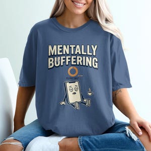 May include: A slate blue short-sleeve t-shirt featuring the text "MENTALLY BUFFERING" in a distressed font above an orange loading circle and the text "PLEASE WAIT...". Below is a cartoon smartphone with tired eyes holding a coffee cup.