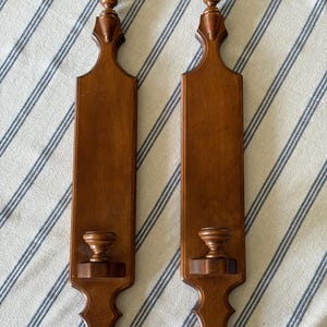 May include: A pair of matching vertical wooden wall sconces with a medium brown finish. Each sconce features a long rectangular body, a decorative finial at the top, a pointed base, and a small circular platform at the bottom for holding a candle.
