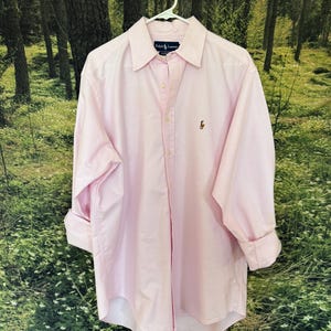 May include: A light pink button-down shirt with long sleeves, hanging on a white hanger. The shirt has a classic collar and a small embroidered logo on the chest. The label reads "Ralph Lauren".
