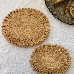 May include: Two oval-shaped, woven placemats. The placemats are made of a light brown, natural fiber. The edges of the placemats have a looped design, creating a decorative border. The placemats are placed on a white surface.