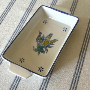 May include: A rectangular white ceramic baking dish with two small handles on the sides. The interior features a central illustration of a yellow corn cob with green husks, surrounded by four small blue floral accents and a blue rim.