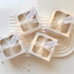 Peut inclure: Quatre boîtes cadeaux blanches avec des fenêtres transparentes, chacune attachée avec de la dentelle blanche et une petite étiquette. Les boîtes sont posées sur une surface blanche texturée avec des perles éparpillées. Les étiquettes indiquent "Emma & Liam" avec une date.