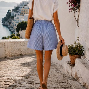 May include: A person walking down a stone path, wearing a white t-shirt, blue and white striped shorts, and brown sandals. They are carrying a straw tote bag and a straw hat with a black band. A coastal town is visible in the background.