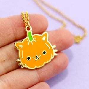 Op de afbeelding: Gouden ketting met een pompoen-vormige hanger met een kattengezicht. De hanger is oranje met een groene steel en zwarte ogen.