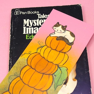 Op de afbeelding: Een roze bladwijzer met een cartoonkat op oranje pompoenen, geplaatst in een vintage boek. Het boek heet "Mystery Image" van Pan Books. De bladwijzer heeft een gradiënt achtergrond.