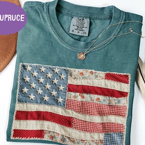 May include: Teal Comfort Colors t-shirt with a sewn-on American flag design. The flag features white stars on a blue background and red and floral-patterned stripes. A gold necklace with a pendant rests on the shirt.