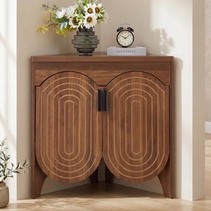 May include: A wooden corner cabinet with two doors featuring arched, concentric oval carvings and vertical black handles. On top sits a dark gray vase with white and yellow flowers, a black alarm clock, and a book labeled CONSIDERED.