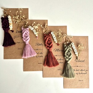 Pode incluir: Quatro porta-chaves em macramé nas cores roxo escuro, rosa claro, terracota e verde sálvia, cada um com fecho dourado, contas de madeira com letras que formam nomes e uma borla. Estão presos a cartões castanhos com flores secas.