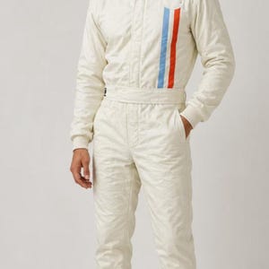 May include: A cream-colored racing jumpsuit featuring a high collar, long sleeves with ribbed cuffs, and a belted waist. The front displays a vertical stripe pattern in light blue and orange, with a circular white patch on the upper left chest.