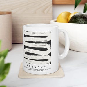 Puede incluir: Una taza de cerámica blanca con un diseño abstracto de pinceladas en blanco y negro. Debajo del gráfico, la palabra PRESENT está impresa en negro, seguida del texto: Being fully in the now. Not dwelling in the past, nor anxious about the future. Simply here.