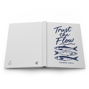 Puede incluir: Un cuaderno blanco abierto con un gráfico azul oscuro en la portada derecha que muestra tres peces apilados verticalmente y el texto Trust the Flow. La parte inferior incluye el texto NUMEN SOUL, mientras que la portada izquierda muestra un pequeño logo y un código QR.