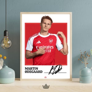 May include: A framed poster featuring a digital illustration of Martin Ødegaard wearing a red and white Arsenal jersey. The design includes the text "MARTIN ØDEGAARD" and a black signature on a white background with a red square behind the player.