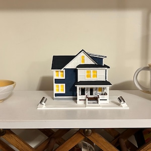 May include: A miniature model of a two-story house with dark blue siding, white trim, and bright yellow windows. The house features a front porch with steps and is positioned on a white rectangular base with two small silver cylindrical accents.