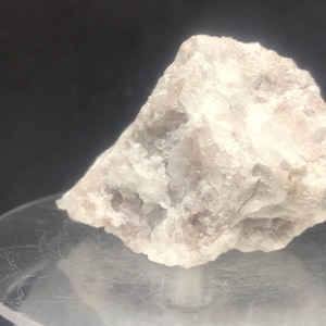 Puede incluir: Primer plano de una formación rocosa rugosa y clara. La roca tiene una estructura cristalina con una mezcla de tonos blancos y rosa pálido. La superficie es texturizada y desigual, con pequeñas cavidades.