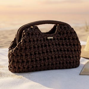 Puede incluir: Un bolso de mano tejido marrón oscuro con asa superior descansa sobre una toalla blanca en una playa de arena. Un sombrero de paja y un libro abierto con las palabras "Beach Reads" también están presentes. El bolso es rectangular.