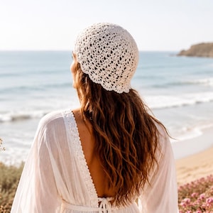 Puede incluir: Una mujer con un sombrero de ganchillo color crema y un vestido blanco, vista de espaldas, mirando una playa y el mar. El vestido tiene la espalda baja. La escena está iluminada por el sol, con una playa de arena y acantilados.