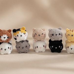 May include: A collection of handmade crochet cat figurines in various colors, including pink, brown, gray, black, yellow, and white. Each cat has a unique design and is stacked on top of another. The figurines are small and appear to be keychains or decorative items.