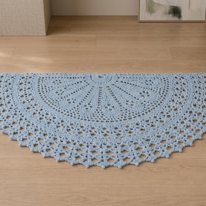 Puede incluir: Una alfombra de ganchillo semicircular de color azul claro con un intrincado patrón de abanico radiante que se extiende desde el centro hasta un borde festoneado calado. La pieza textil muestra una textura de punto consistente.