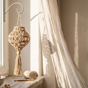 May include: A beige crocheted lantern with an openwork pattern, hanging from a metal wall hook. The lantern features a braided cord handle, a wooden bead at the base, and a long, dangling tassel. The design is crafted from light-colored yarn.