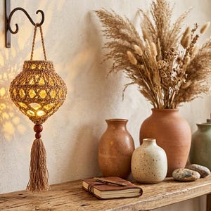 May include: A hanging lantern crafted from tan woven cord with a geometric pattern, featuring a central light source, a dark wooden bead, and a long matching tassel. The lantern is suspended from a black metal wall hook, casting a warm glow.